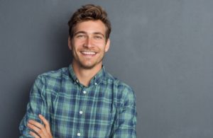 Young handsome man leaning against grey wall with arms crossed. Cheerful man laughing and looking at camera with a big grin. Portrait of a happy young man standing with crossed arms over grey background.